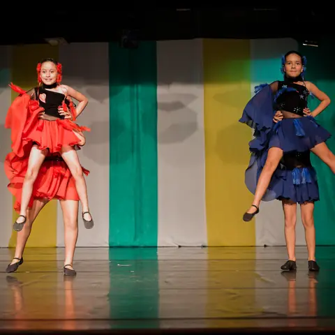 Duas bailarinas de saia vermelha e azul no meio de um salto.