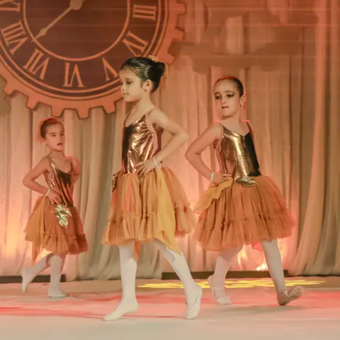 Bailarinas de dourado diante do relógio do cenário.