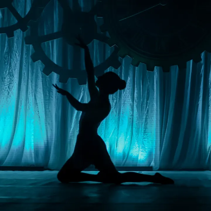 Silhueta de uma bailarina ajoelhada, com o braço estendido para o alto, contra a cortina iluminada de azul.